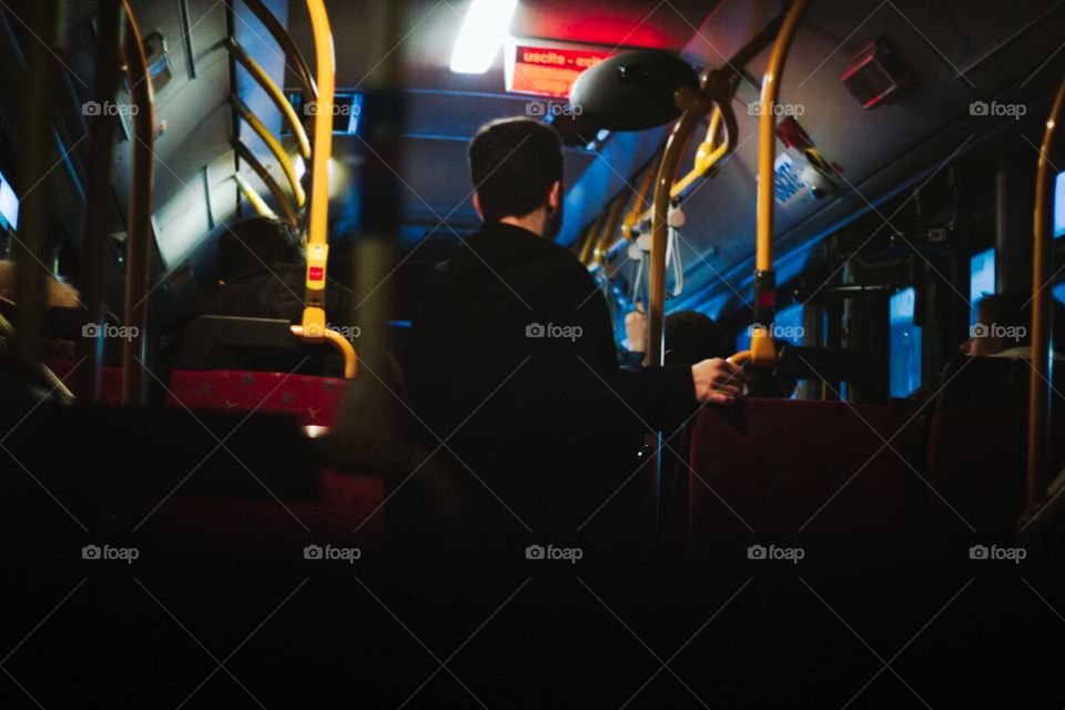 Urban photo of a man on the bus waiting for the next bus stop to go home, overhead lighting and a blue cast from the sky outside 1 hour before sunset
