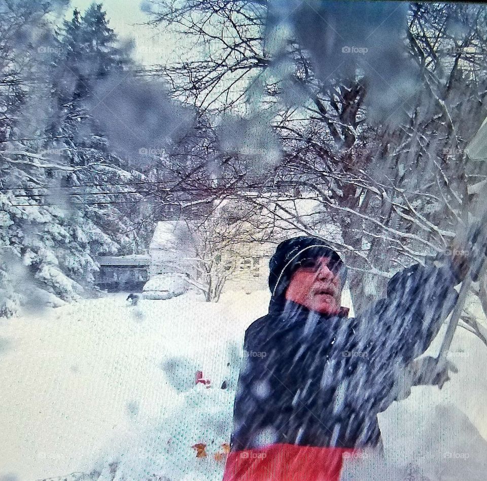 Brushing snow off gutters before freeze, dressed for snowstorm as man shovels. Snow cap, sunglasses, gloves, ski jacket on.