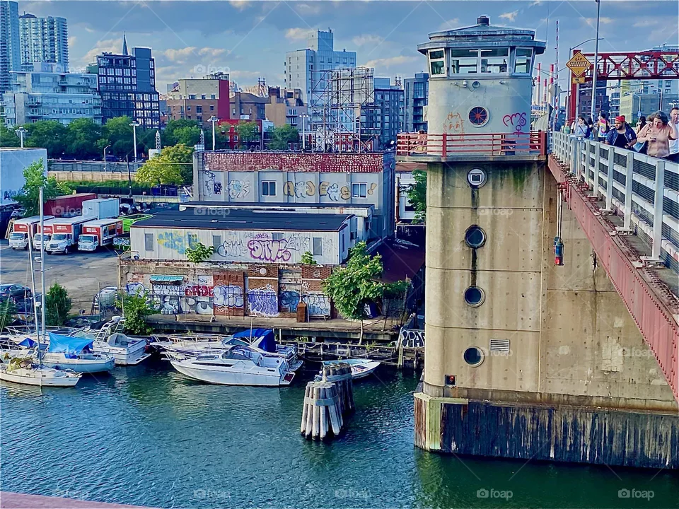 The “Pulaski Bridge” featuring one of its “watchtowers” and main bridge supports with a view towards “LIC, Queens” with some colorful graffiti, green vegetation in between and boats by the shore. 2022. Hypnotic Productions