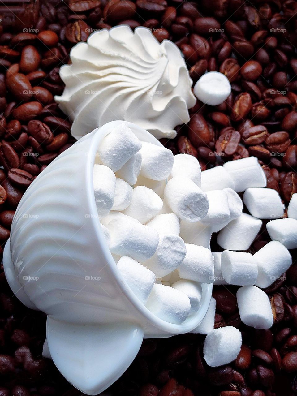 White colour.  On brown coffee beans lies an overturned white cup with a white marshmallow that has spilled out of the cup.  In the background lies a white marshmallow in the shape of a seashell.  Contrast