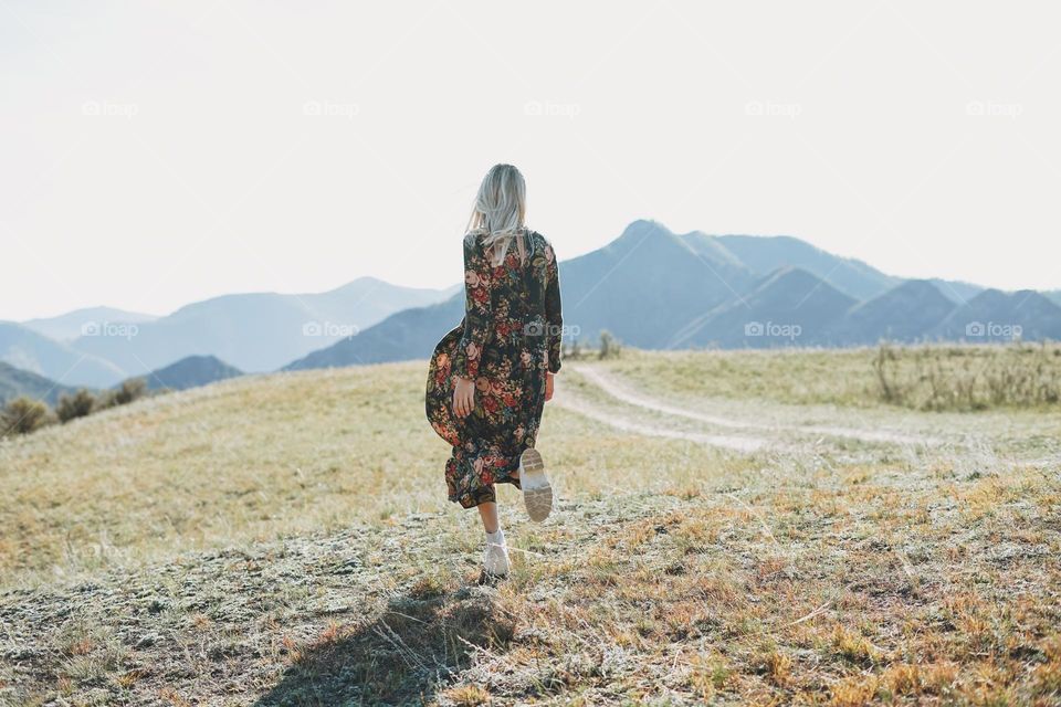 Beautiful blonde young woman traveler in dress from back on road, trip to mountains