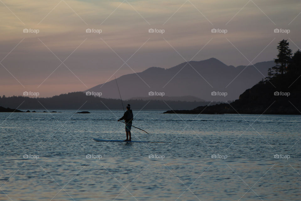 The sun has set behind the mountains & a woman paddling out in the cove to catch a fish or two can be seen silhouetted against the inky blue, calm water. The dark rocky outcrops & purple mountains are silhouetted against the pink & purple sky. 