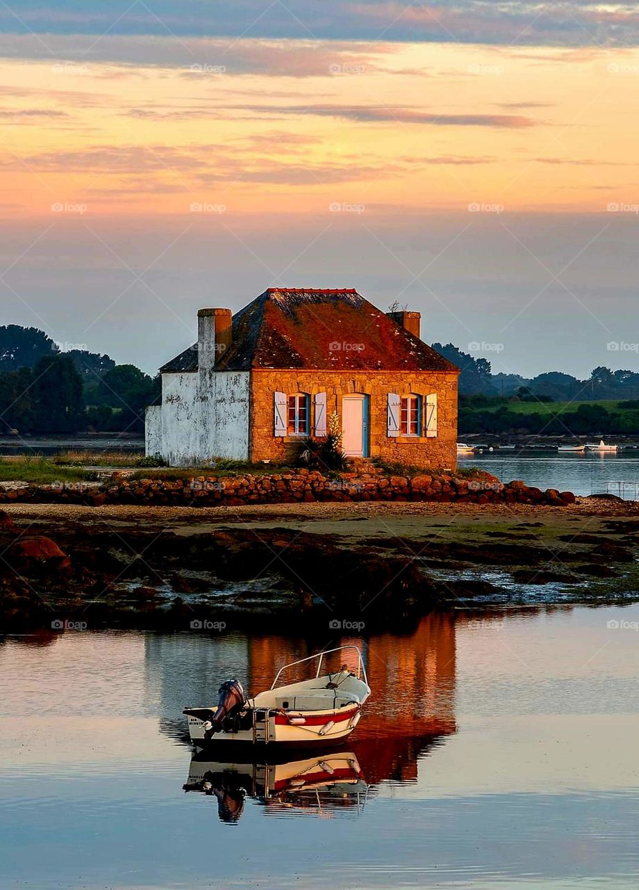 The only building on Saint Cado Island, a small house with blue shutters on the rocks of Nichtarguer and a fisherman's boat both reflecting in the sea at sunset