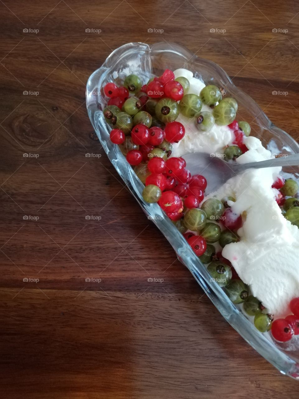 In the elongated see-through glass bowl there are a little melted vanilla ice-cream sundae with plenty of red and green berries and a teaspoon, made of steel. The wooden brown table with knots.