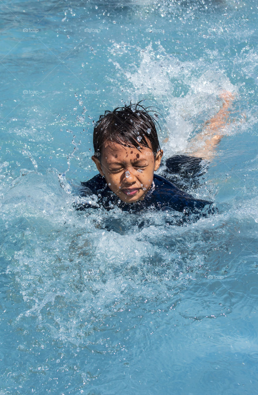seorang anak kecil yang sedang belajar berenang disebuah kolam dangkal dengan air yang berwarna biru. anak kecil tersebut tampak memejamkan mata karena percikan air yang mengarah ke matanya.