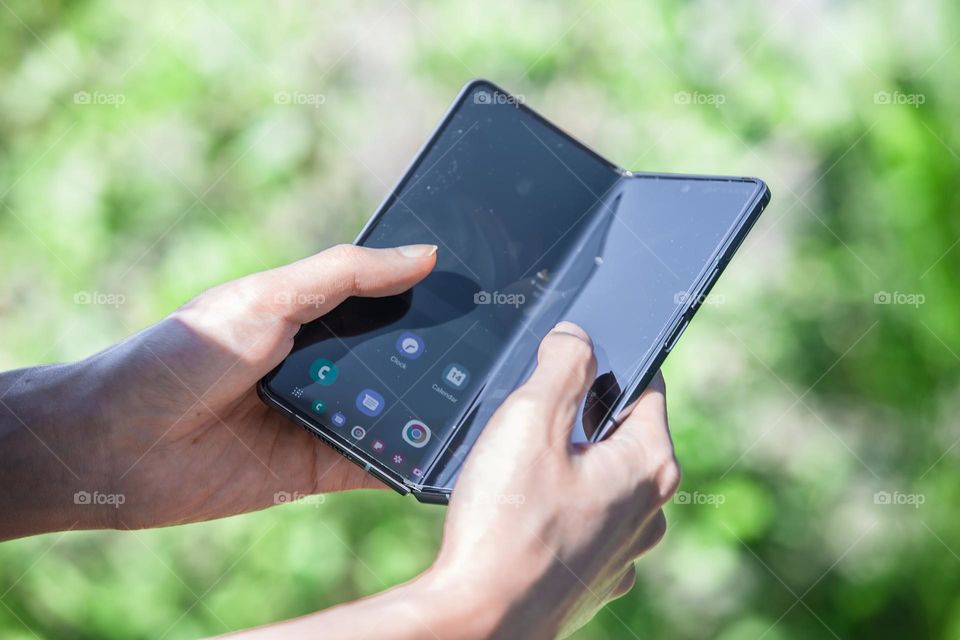 All new Samsung Galaxy Z Fold 4 in gray green color. An Asian man uses it outside in the park. This is the most recent foldable phone released by the Korean company. Greenery from surrounding leaves in bokeh in background.