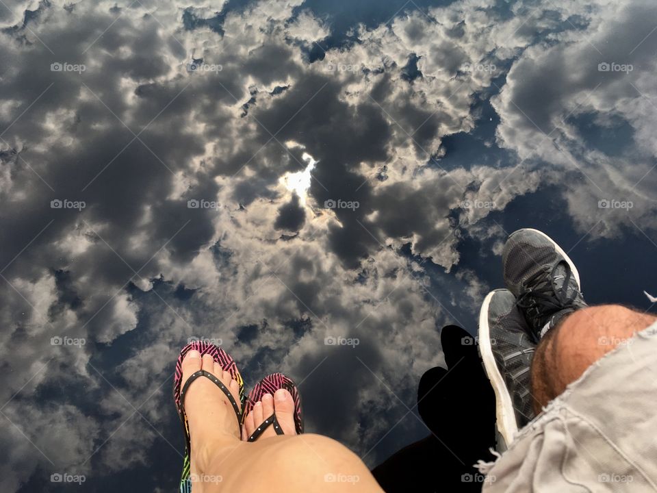 Legs hanging over water with a reflection of the sun in a cloudy sky