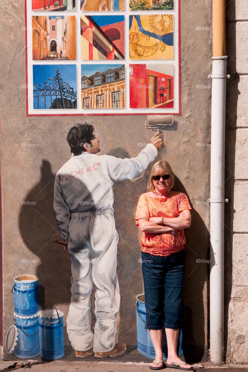 Woman leans on the wall next to a mural of a man painting the wall.