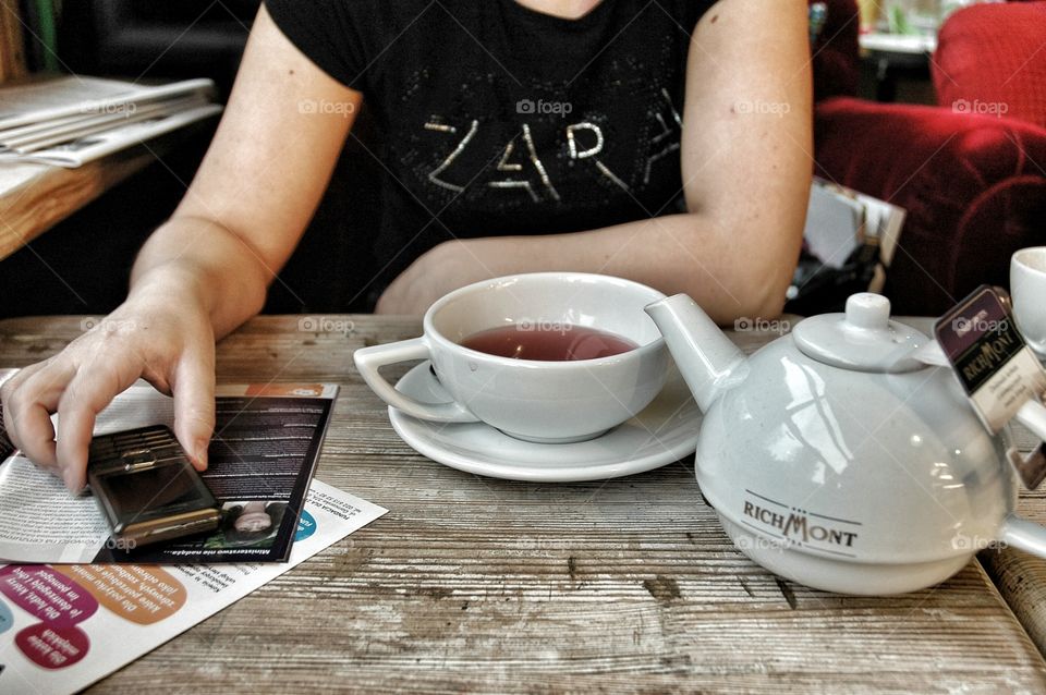 Young woman at cafe sitting at table with cup of tea in front of her using mobile phone