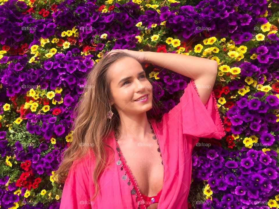 Portrait of pretty young woman wearing pink dress surrounded by wall of multicoloured flowers