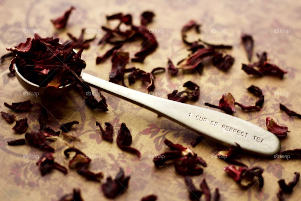 Angled view of a stainless steel measuring spoon imprinted with the phrase, “1 CUP OF PERFECT TEA,” filled with dried hibiscus petals on a brown and burgundy patterned surface with scattered hibiscus tea