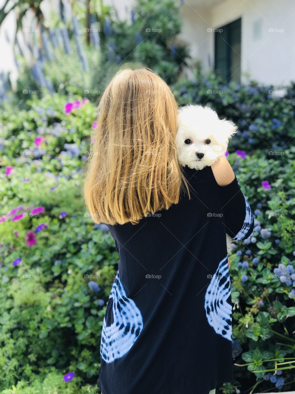 Puppy peeking over shoulder of blond girl who is holding puppy lovingly with flowers and greenery all around them. 