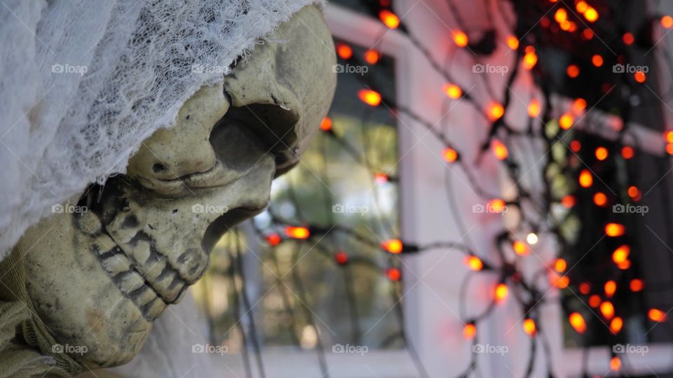 A skeleton in a shroud, on a porch with holiday lights in the background.