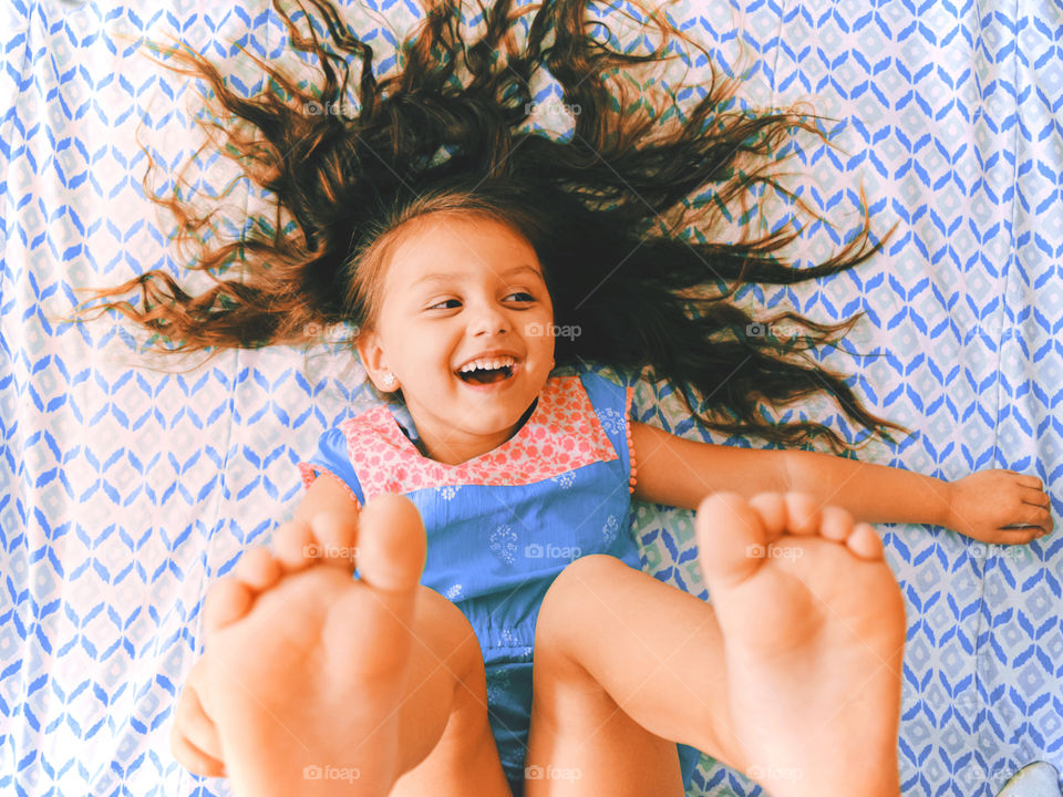 A view from the top on a happy little girl smiling lying on the bed and showing her feet