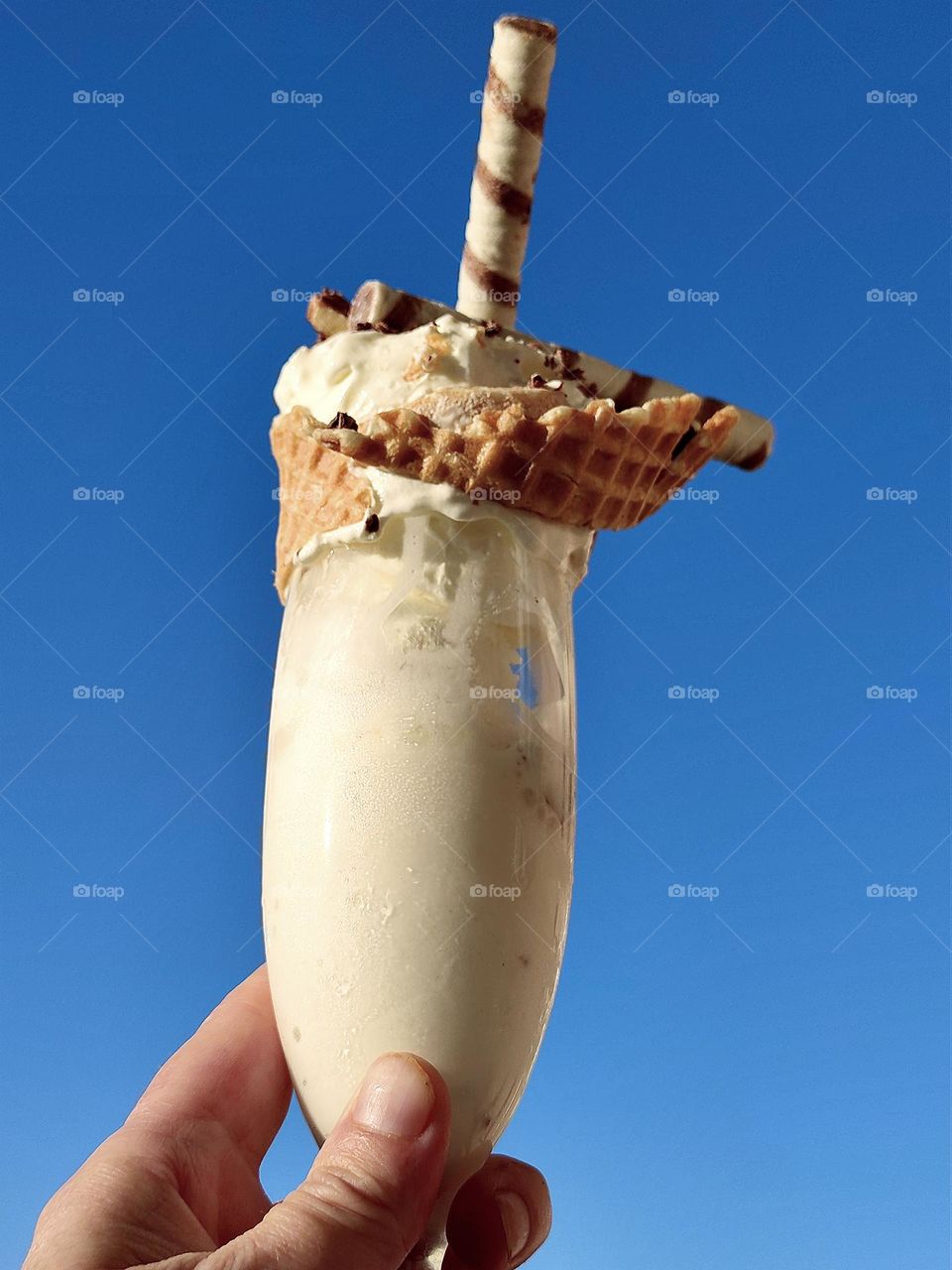 White color. A woman's hand holds a glass of white ice cream against a blue sky. The edge of the glass with ice cream is wrapped with a faffle sheet. Ice cream sprinkled with grated chocolate, nuts and waffle sticks