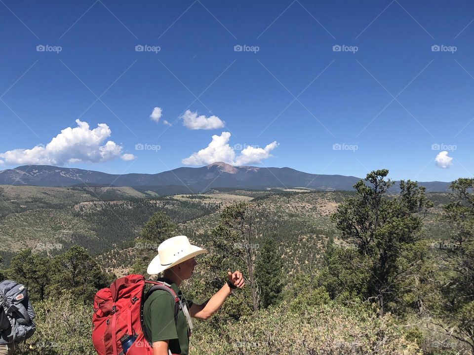 Backpacking cowboy stands before mountainous background