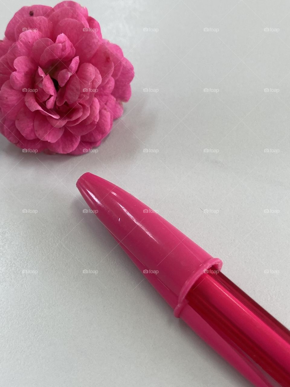 Caneta pink apontando uma rosa pink, ambos objetos estão sobre uma mesa cinza clara e lisa.