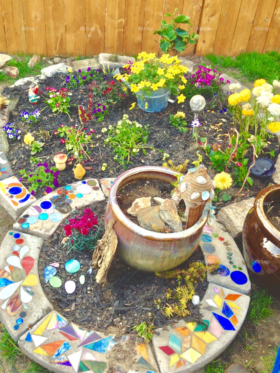 "Fairy Garden" with different plants and colorful flowers, surrounded by rocks, seashells, fairy figurines, colorful stones and peices of slate, all arranged creatively in designed circular enclosures, some transplanted from pots, some still in pots.