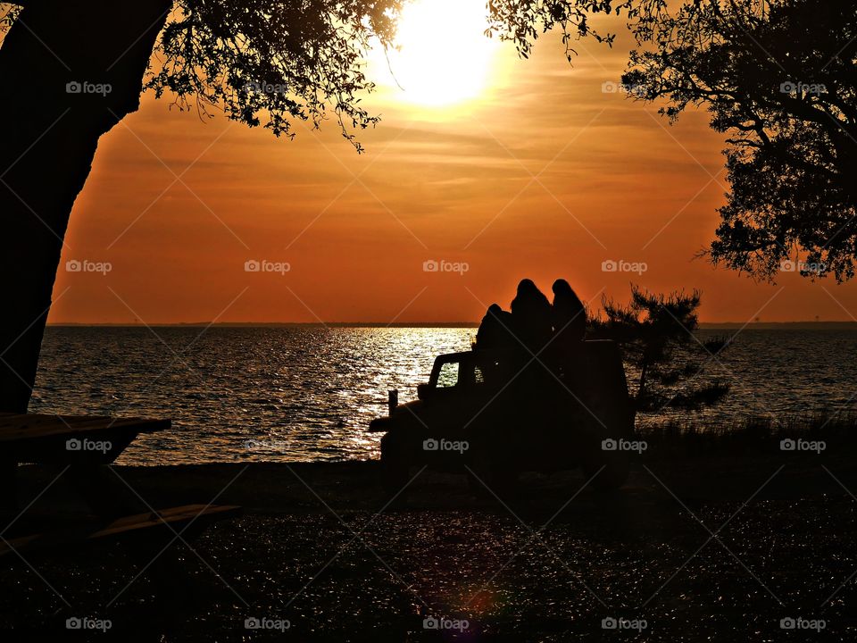 
Silhouette of three adult people sitting on top of a Jeep talking and looking at the colorful sunset 