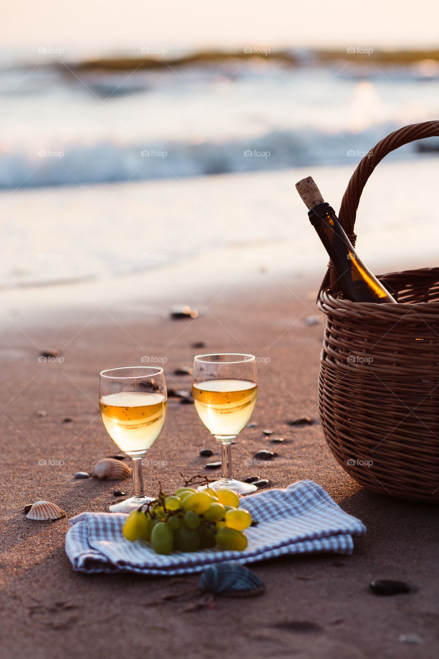 Two wine glasses with white wine standing on sand, on beach, beside grapes and wicker basket with bottle of wine. Sea waves in the background