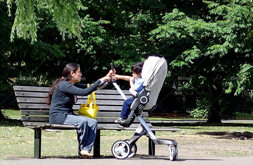 A mother with expressions of pure joy as she sits on a bench with her son in a stroller on a walk in Regents Park on a sunny summer day. 