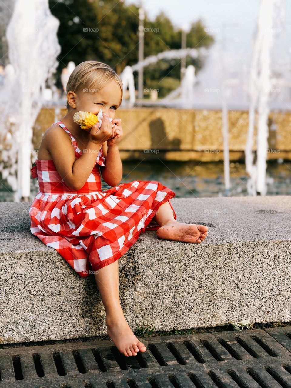 Cute little girl with short blonde hair wearing red dress sitting near fountains and eating corn, portrait of child 