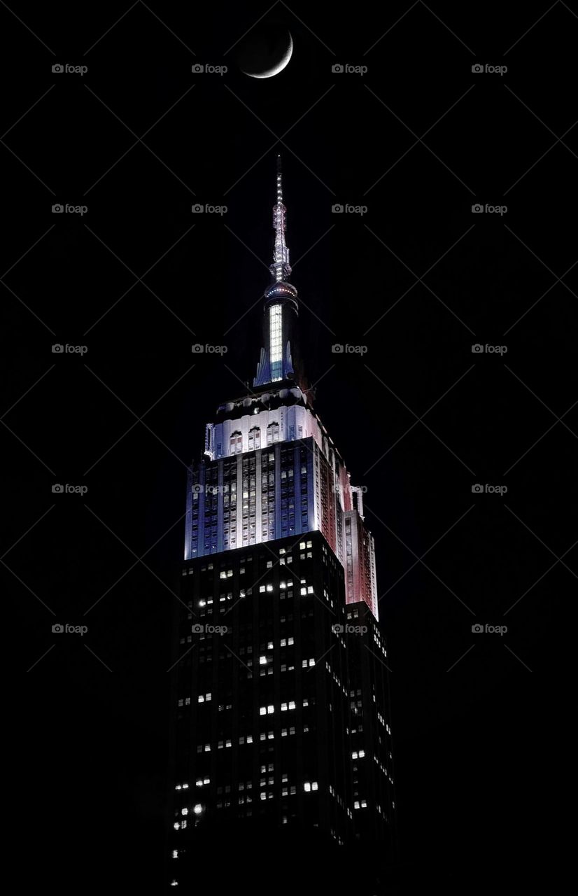 The Empire State Building lit in red, white and blue lights at night with a crescent moon overhead 