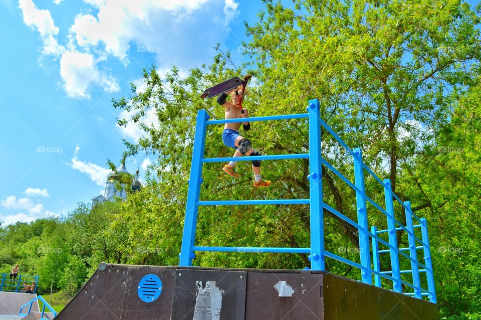 a young man of athletic build is in the air performing a trick on a stunt scooter. green trees and blue summer sky in the background.