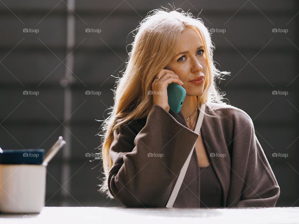 Young beautiful blonde woman sitting in modern cafe and talking on the phone in sunny day, portrait of woman 