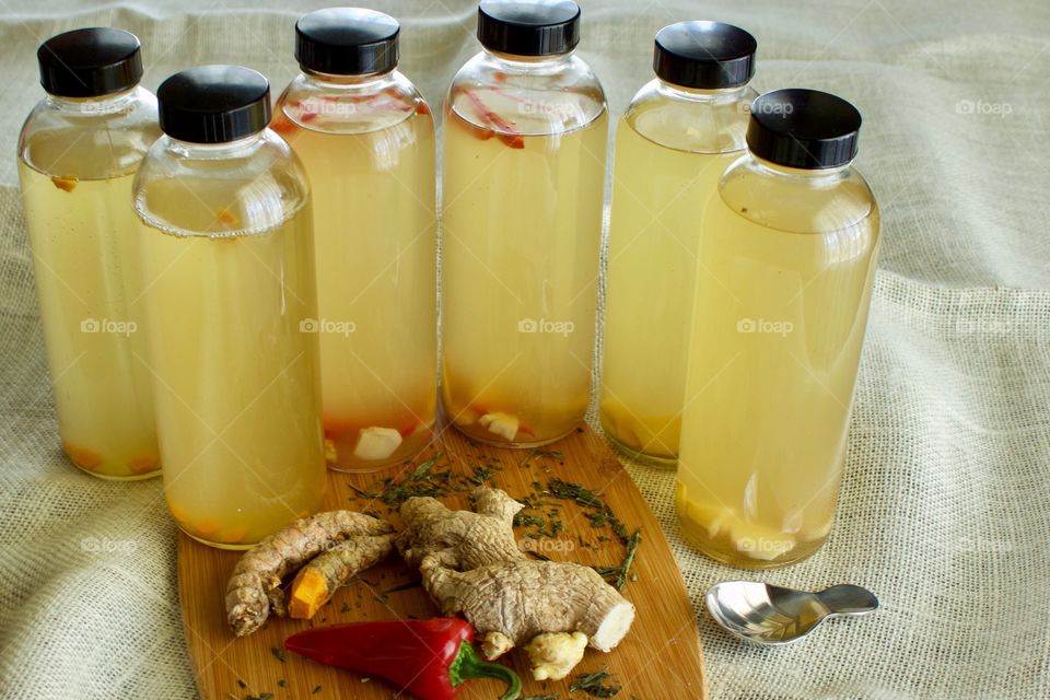 Homemade kombucha, bottled for a second ferment, flavored with slices of turmeric root, red chili pepper, and / or ginger root, with scattered, dried bancha leaf tea, and stainless steel tea scoop on bamboo cutting board