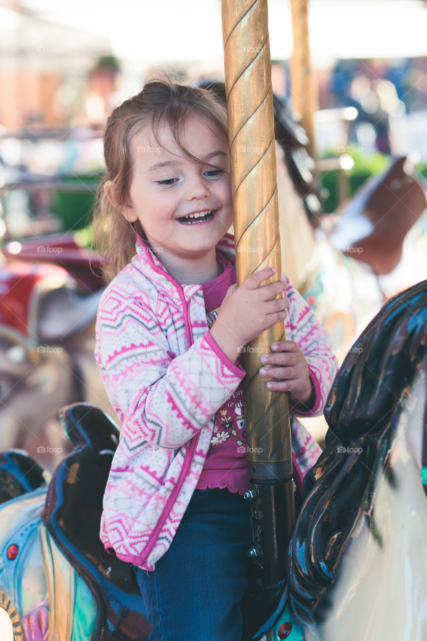 Little adorable smiling girl riding a horse on roundabout carousel at funfair