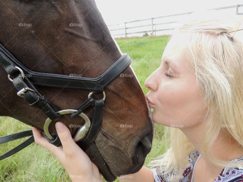 Blonde girl kissing the nose of her dark bay horse wearing an English bridle in a field.