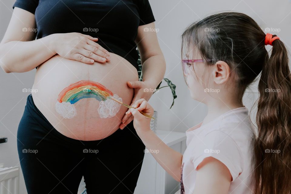 Portrait
little caucasian girl painting a rainbow with clouds on her pregnant mother's belly with a brush and acrylic paint, close-up side view.