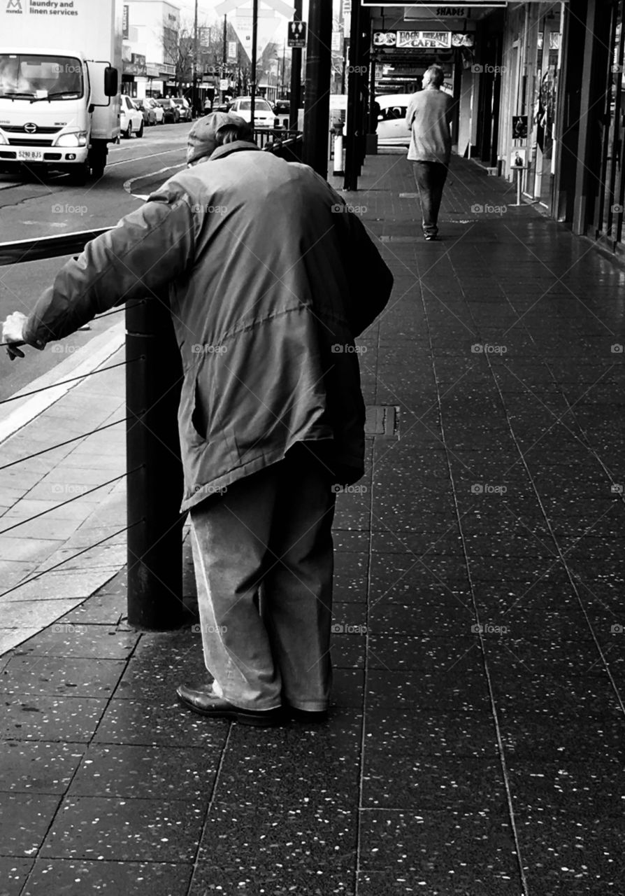 Black and white image poor elderly man hunched wearing coat on city street 