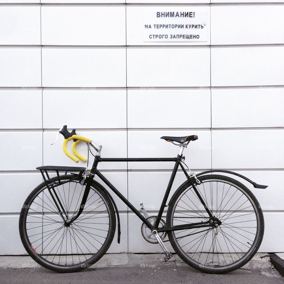 Black singlespeed soviet commuter bicycle with a front bicycle rack and yellow handlebar tape in front of white wall in Moscow, Russia