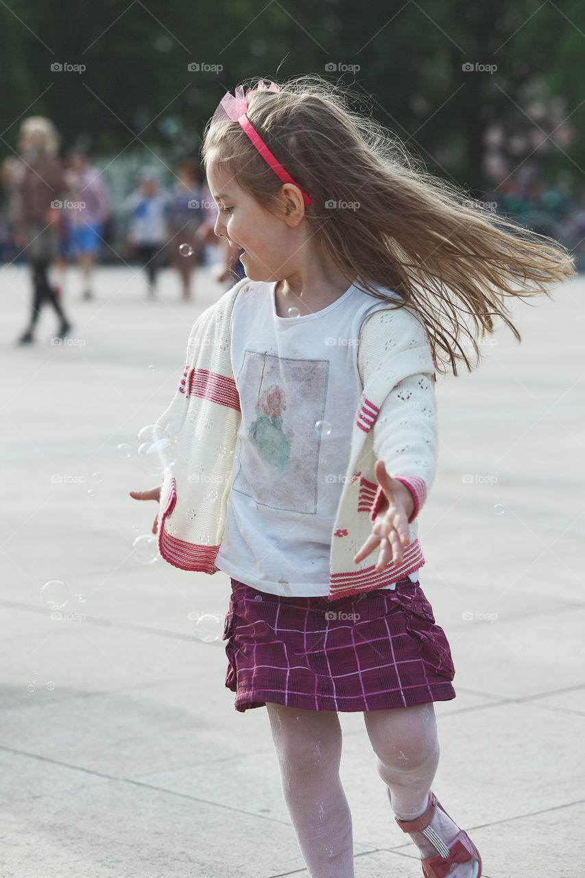 Little happy girl playing with the soap bubbles in the center of town. Child running around and catching the soap bubbles. Candid people, real moments, authentic situations