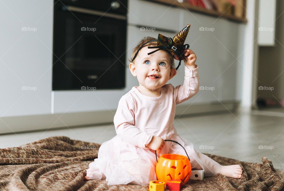 Cute little baby girl in pink dress with the witch's hat and pumpkin lantern sitting on floor in kitchen at the home, Halloween time
