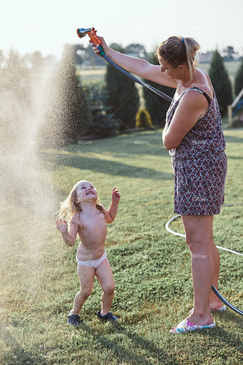 Little cute adorable girl enjoying a cool water sprayed by her mother during hot summer day in backyard. Candid people, real moments, authentic situations