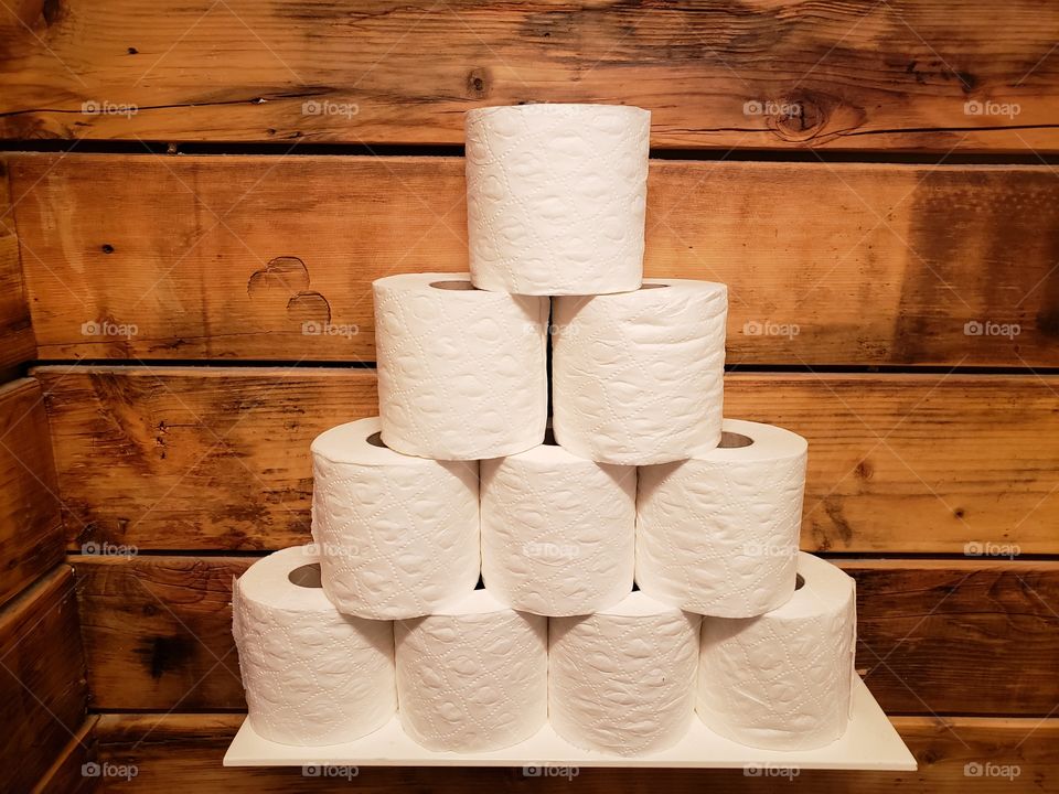 A pyramid of ten stocked rolls of toilet paper is placed in front of a rustic wooden wall in a bathroom or closet