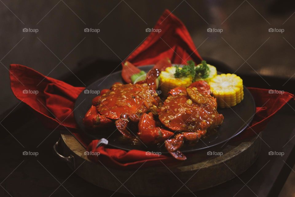 Crab flavored in creamy garlic sauce served with corn, tomatoes, lemon wedges and cherries on a black plate set on a red cloth set in a wooden setting. Asian flavor crab.