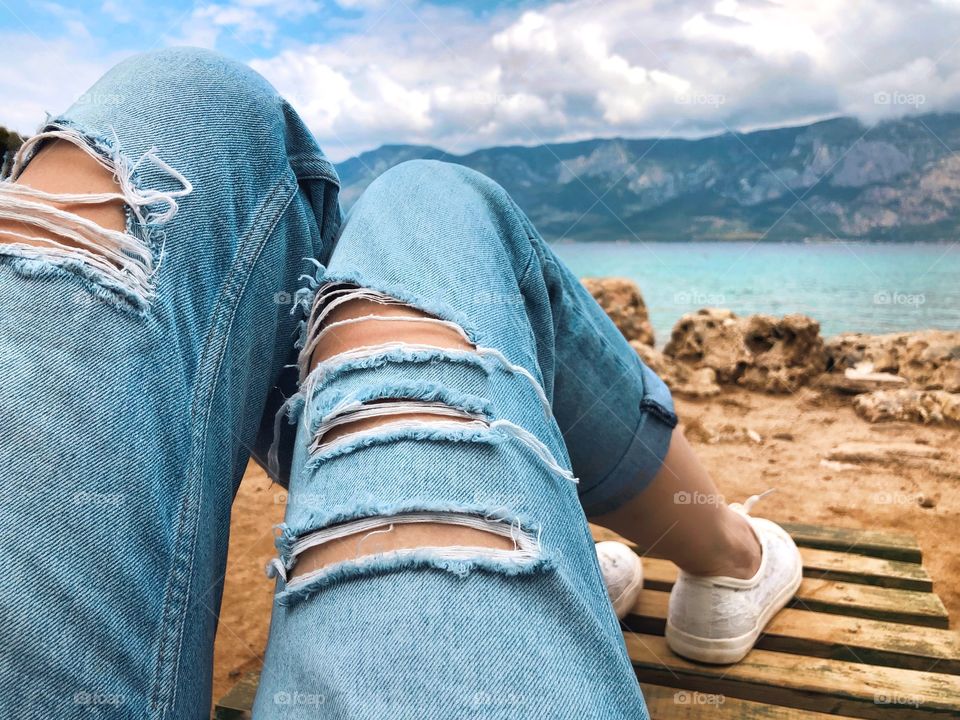 View of the legs of a girl in ripped jeans on a background of a lake and mountains