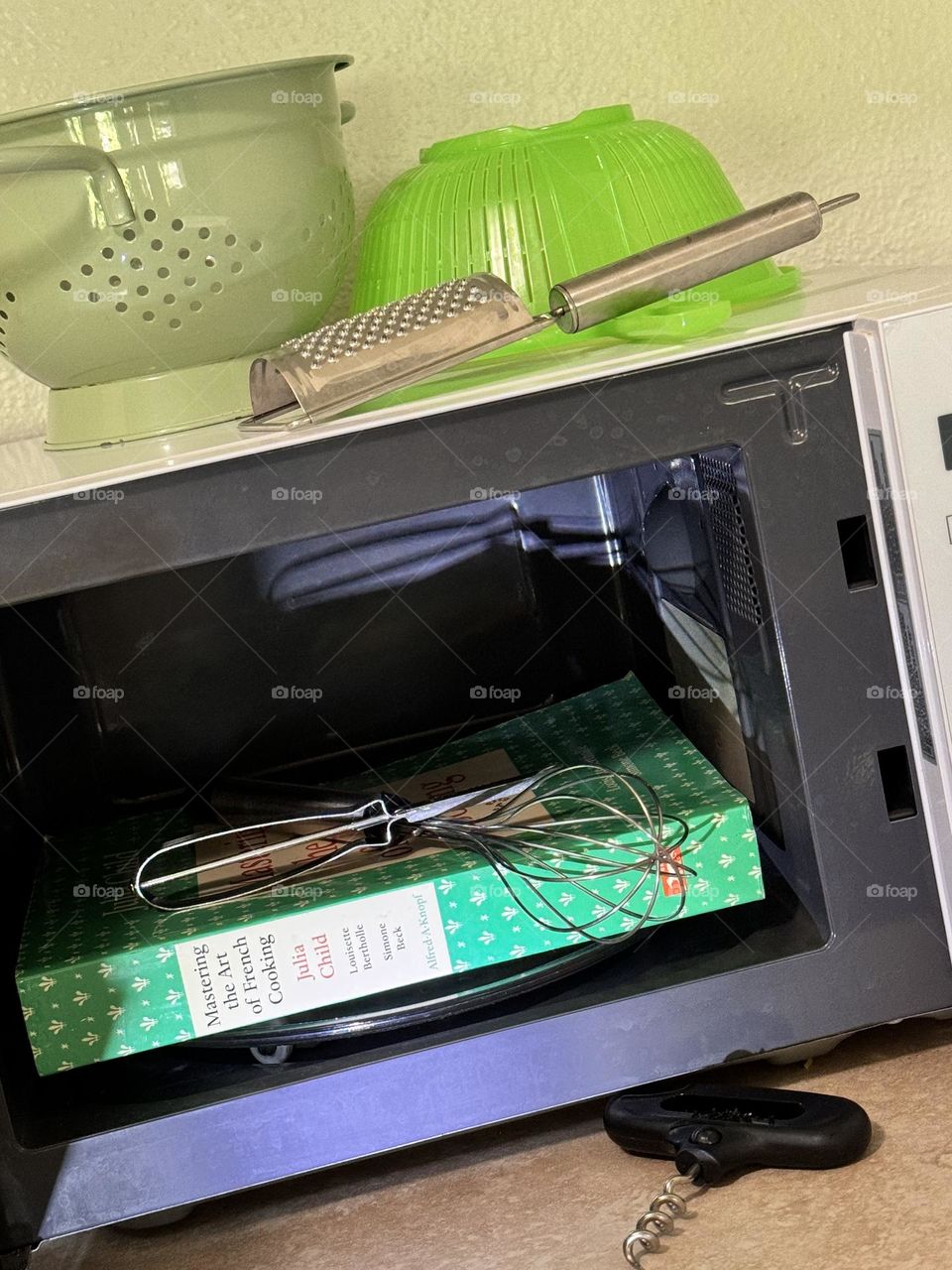 Julia Child’s Mastering the Art of French Oooking cookbook inside microwave oven with corkscrew potato peeler whisk cheese grater and two sieves