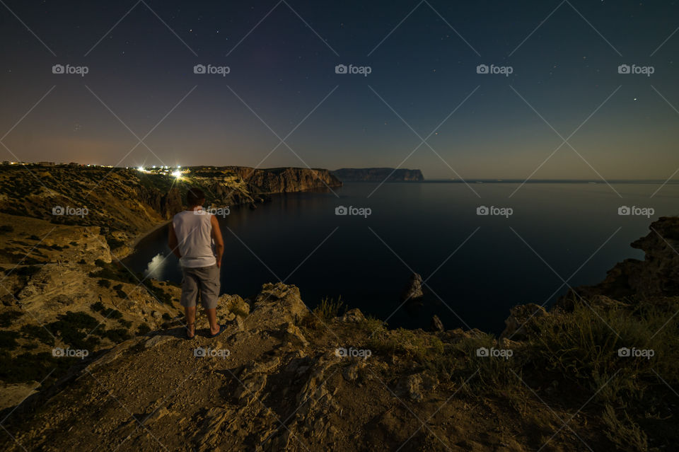Cape Fiolent, in the foreground, on the edge of a cliff stands a young man and admires the nightly Black Sea landscape