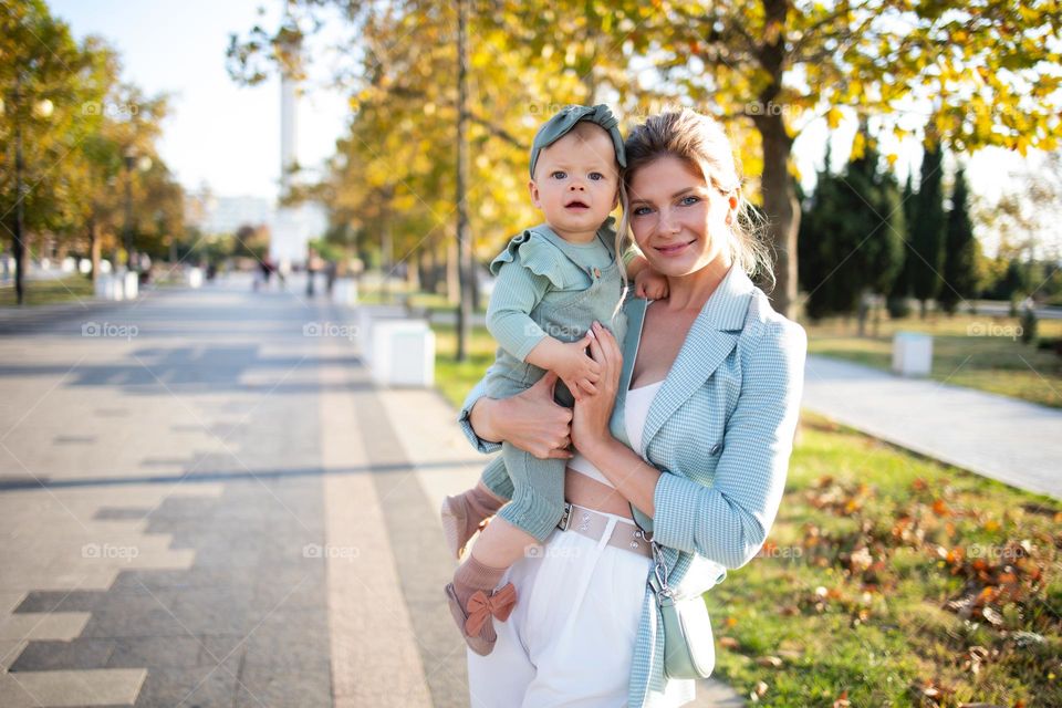 Mother holds her little daughter in her arms among in the autumn park. Mom stylish in mint jacket and mint knitted jacket and her little baby. Concept: child care, children's happiness, adoption. Child up to one year