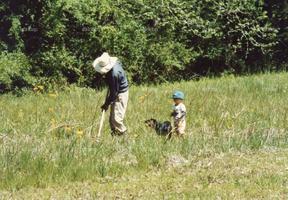 Grandfather working in the pasture during the day with his preschool grandson  and his dog watching.