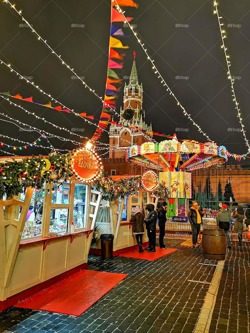 Red Square in a festive decoration. Tents with Christmas decorations and food. Bright lighting. Spirit of Christmas. People are standing at the Christmas tents. In the background the Kremlin's Spasskaya Tower