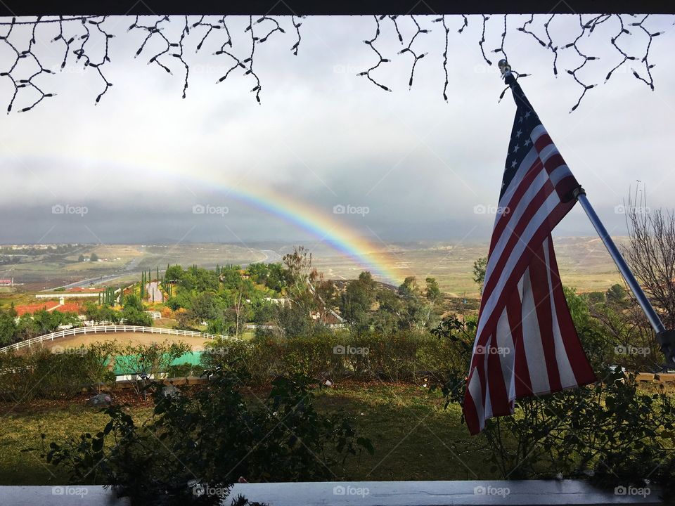 This photo captures a beautiful scene in wine country. Christmas lights hang from the porch with an American flag on the right looking out to a rainbow and dramatic clouds.