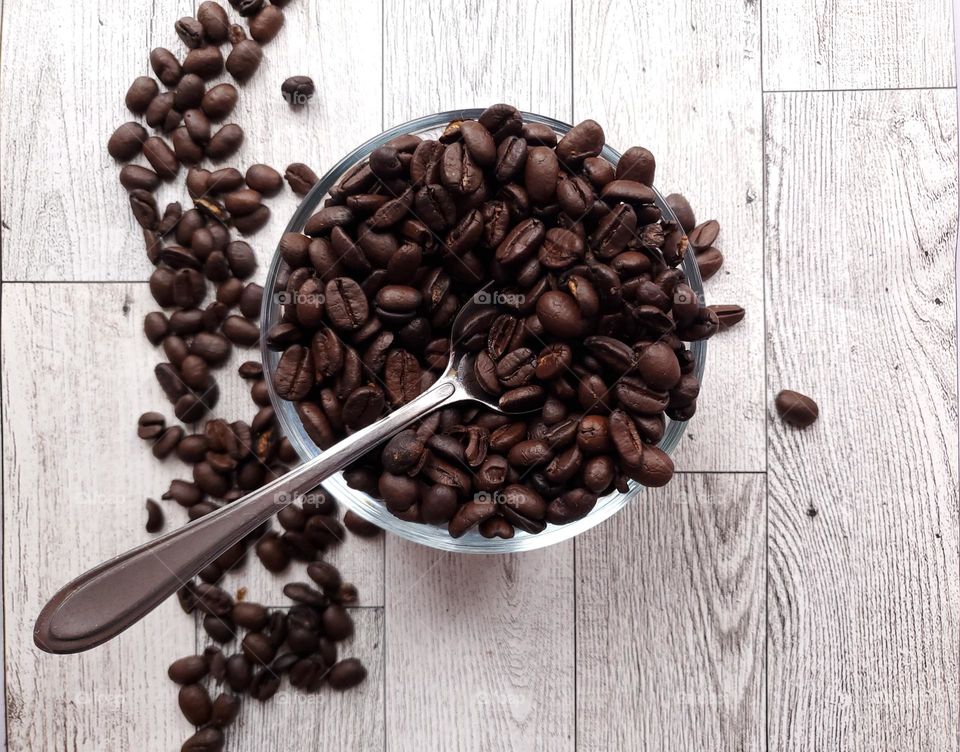 A glass bowl of coffee beans with a spoon and accent coffee beans on the left. Light colored vertical wood surface background.