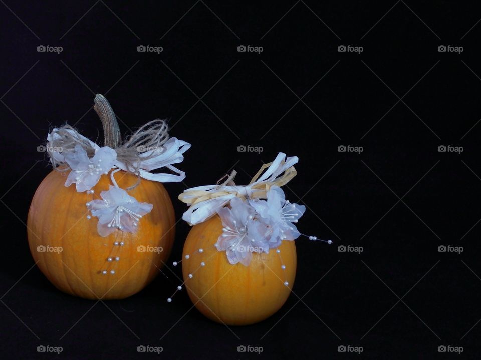 Two small pumpkins decorated country style for a wedding with light brown and white paper ribbons and white flowers and beads all against a black background with ample copy space. 