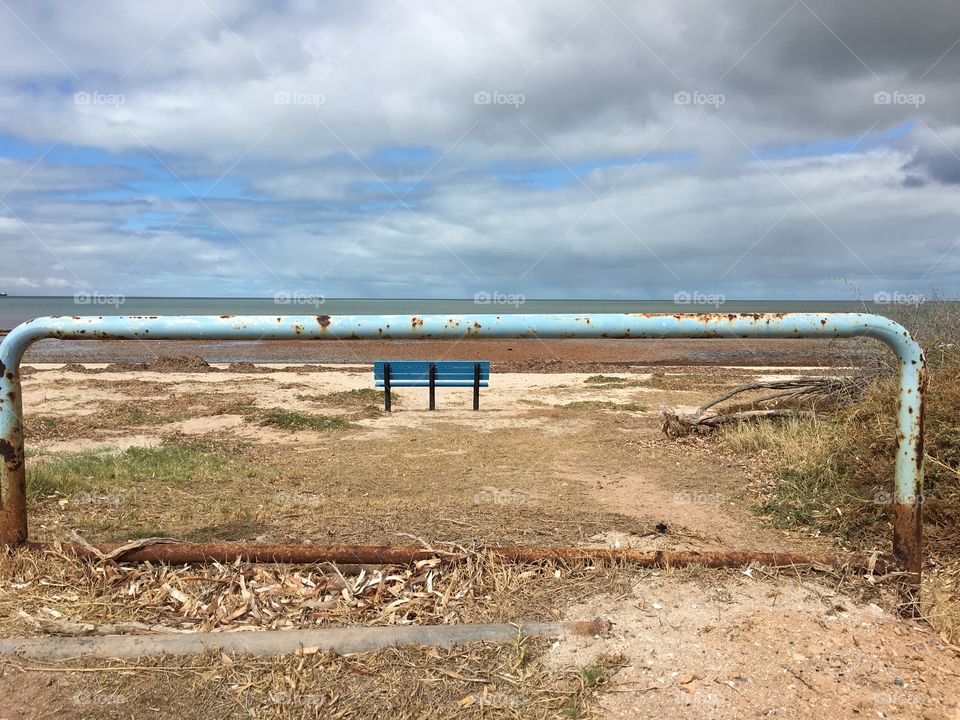 Rusty metal aqua marine blue pole barrier with blue bench, sandy beach, and ocean beyond on billowy cloudy day